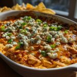 Close-up of a baked buffalo chicken dip in a white oval dish, topped with crumbled blue cheese and chopped green onions.