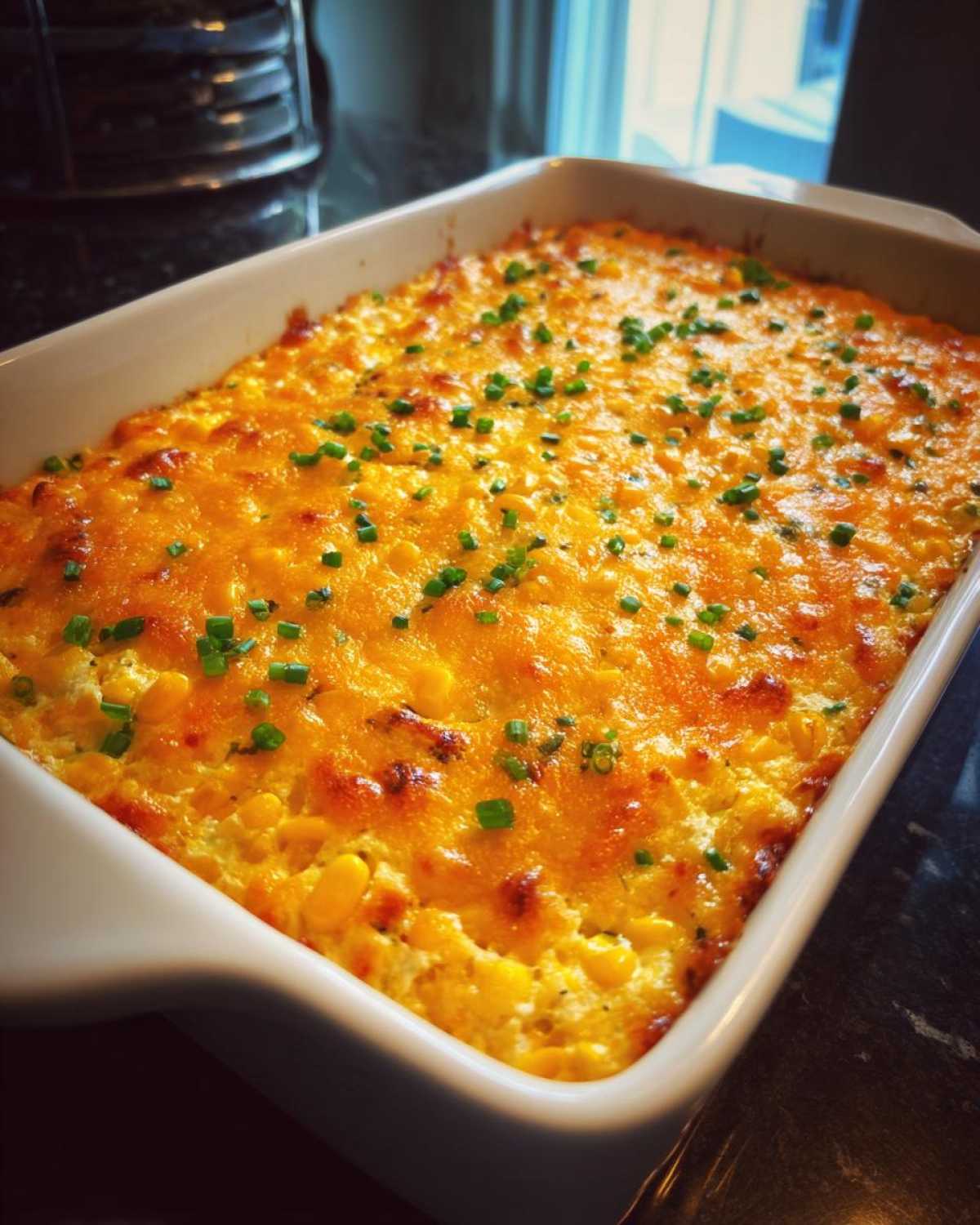 A large white rectangular casserole dish filled with baked corn dip with cream cheese, topped with melted golden cheese and fresh chives.