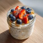 Close-up of overnight oats in a glass jar topped with fresh strawberries, blueberries, chia seeds, and a drizzle of honey.