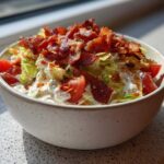 A close-up of a bowl filled with creamy BLT dip, topped with crispy bacon bits, chopped lettuce, and diced tomatoes.