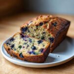 A golden-brown loaf of blueberry zucchini bread with one slice cut and placed in front, showing blueberries and green zucchini pieces.