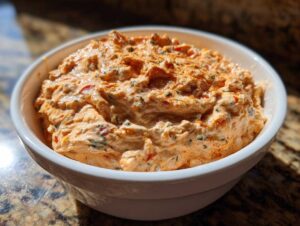 A close-up of a white bowl filled with creamy orange Boat dip Rotel ranch dip, garnished with herbs and spices.