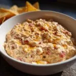 A white bowl filled with creamy Boat dip, topped with melted cheese and black pepper, with tortilla chips in the background.