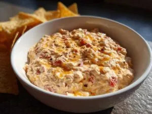 A white bowl filled with creamy Boat dip, topped with melted cheese and black pepper, with tortilla chips in the background.