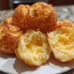 A close-up of golden-brown Brazilian cheese bread (Pão de Queijo) on a white plate, with one cut open revealing its soft, cheesy interior.