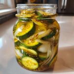 A clear glass jar filled with homemade bread and butter pickles, showing sliced cucumbers, onions, and mustard seeds.