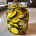 A clear glass jar filled with homemade bread and butter pickles, showing sliced cucumbers, onions, and mustard seeds.