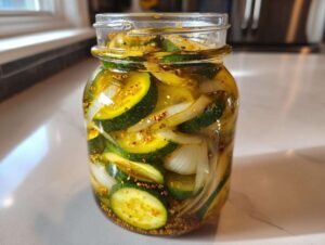 A clear glass jar filled with homemade bread and butter pickles, showing sliced cucumbers, onions, and mustard seeds.