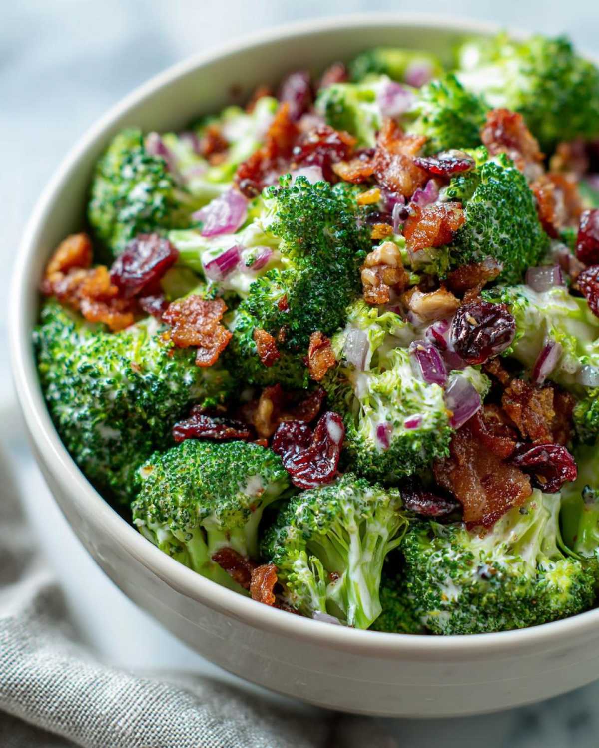 Close-up of a bowl of vibrant broccoli salad with florets, crispy bacon, red onions, dried cranberries, and a creamy dressing.