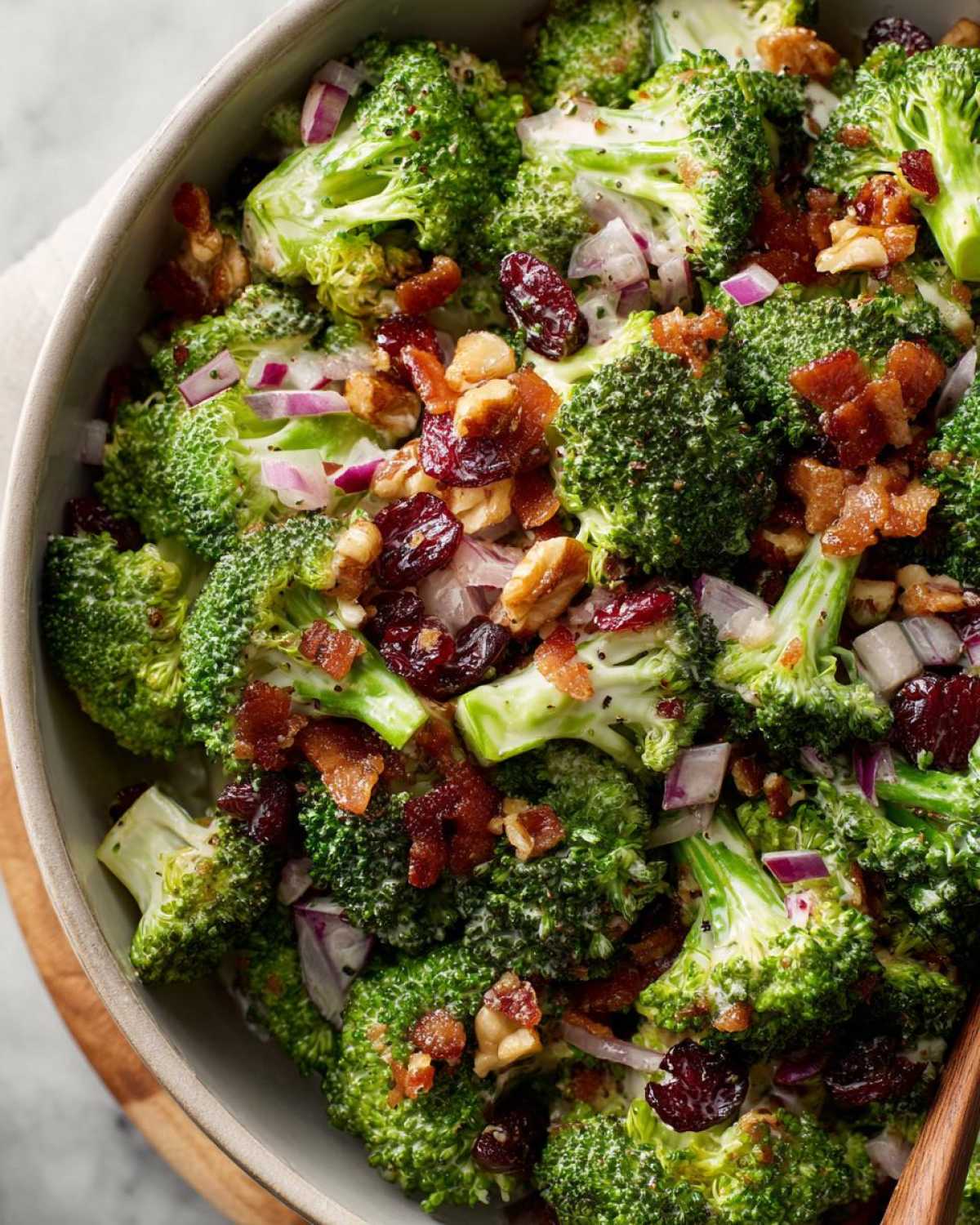 Close-up of a vibrant broccoli salad in a bowl, featuring green broccoli florets, red onions, dried cranberries, and crumbled bacon.