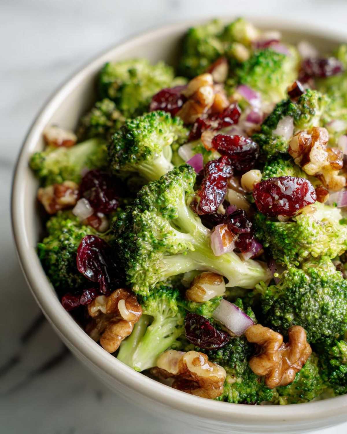 A close-up shot of a vibrant broccoli salad in a white bowl, featuring green broccoli florets, red dried cranberries, and walnuts.