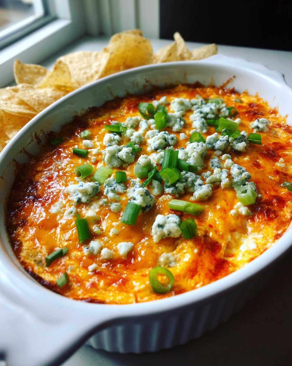 A white baking dish filled with hot, bubbling buffalo chicken dip, topped with blue cheese crumbles and chopped green onions. Tortilla chips are visible in the background.