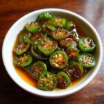 A white bowl filled with vibrant green candied jalapenos, glistening in a sweet, amber syrup with visible seeds and chili flakes.