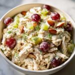Close-up of a vibrant chicken salad recipe in a white bowl, featuring shredded chicken, red grapes, green grapes, almonds, and fresh dill.