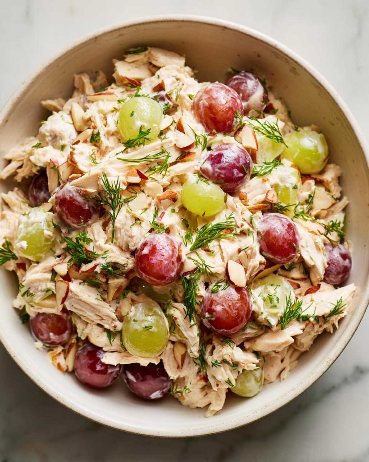 Overhead close-up of a bowl of chicken salad recipe, featuring shredded chicken, red and green grapes, sliced almonds, and fresh dill.
