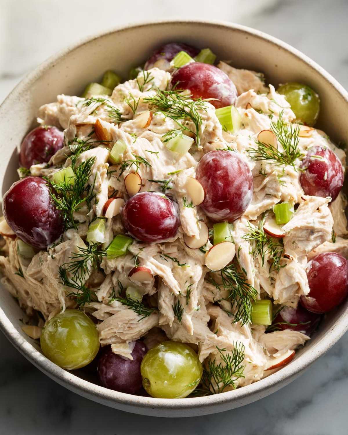 Close-up of a bowl filled with creamy chicken salad, featuring shredded chicken, red and green grapes, celery, sliced almonds, and fresh dill.