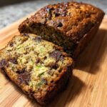 A golden brown loaf of chocolate chip zucchini bread on a wooden cutting board with one slice cut and visible.