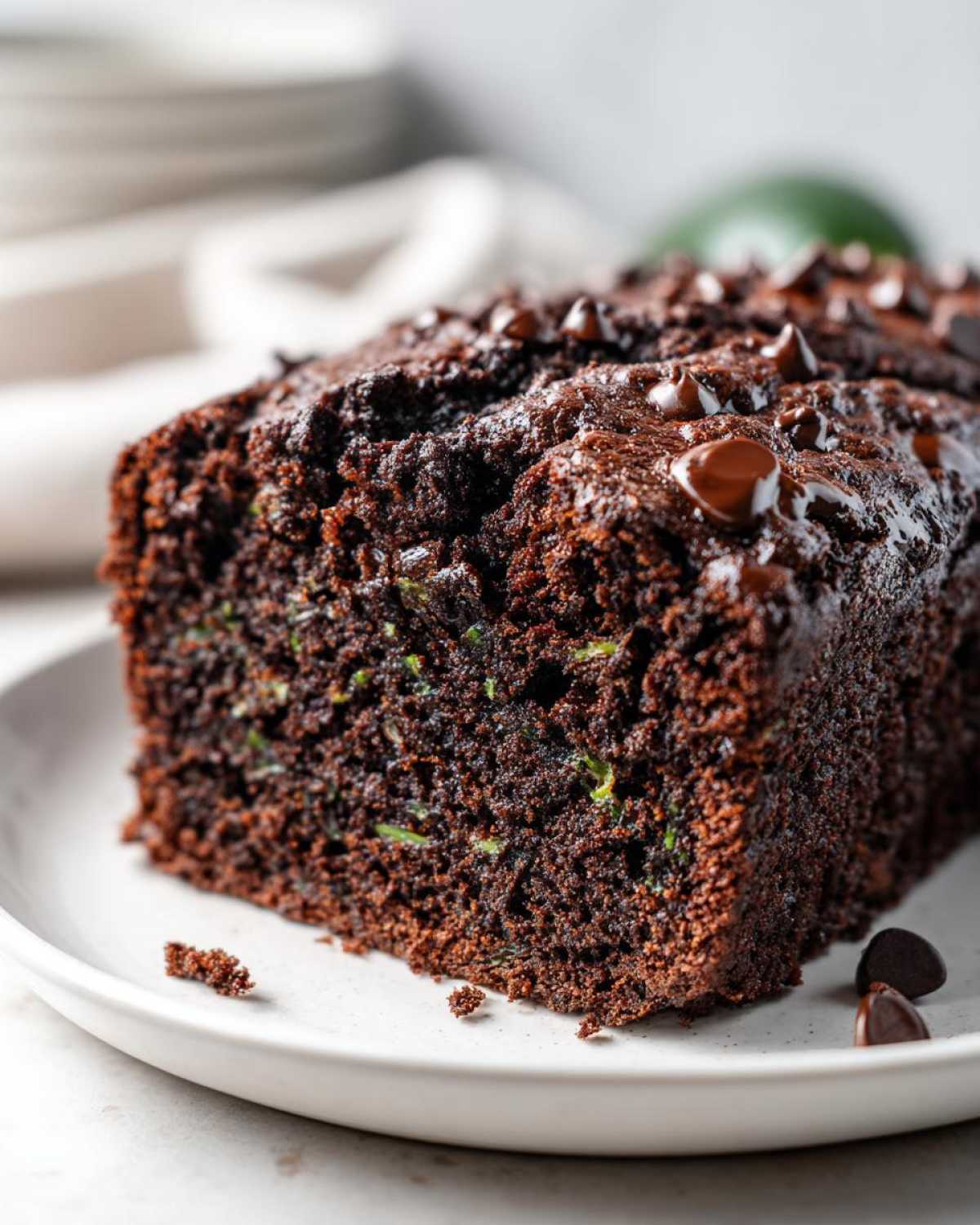 A close-up shot of a rich, dark chocolate zucchini bread loaf on a white plate, topped with chocolate chips.