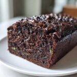 Close-up of a dark chocolate zucchini bread loaf on a white plate, topped with chocolate chips.