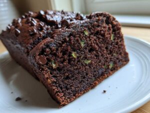A close-up of a dark chocolate zucchini bread loaf with a slice cut, showing green zucchini bits and chocolate chips.