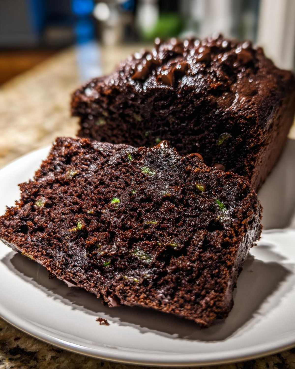 Close-up of a dark chocolate zucchini bread slice on a white plate, showing green zucchini specks and chocolate chips.