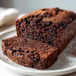 A close-up of a dark chocolate zucchini cake loaf with one slice cut, showing its moist texture and a dusting of cocoa powder.