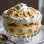 A large glass trifle bowl filled with layers of creamy banana pudding, sliced bananas, vanilla wafers, and topped with whipped cream and a sprinkle of cinnamon.