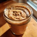 Close-up of creamy blended overnight oats in a glass mason jar on a wooden surface, illuminated by natural light.