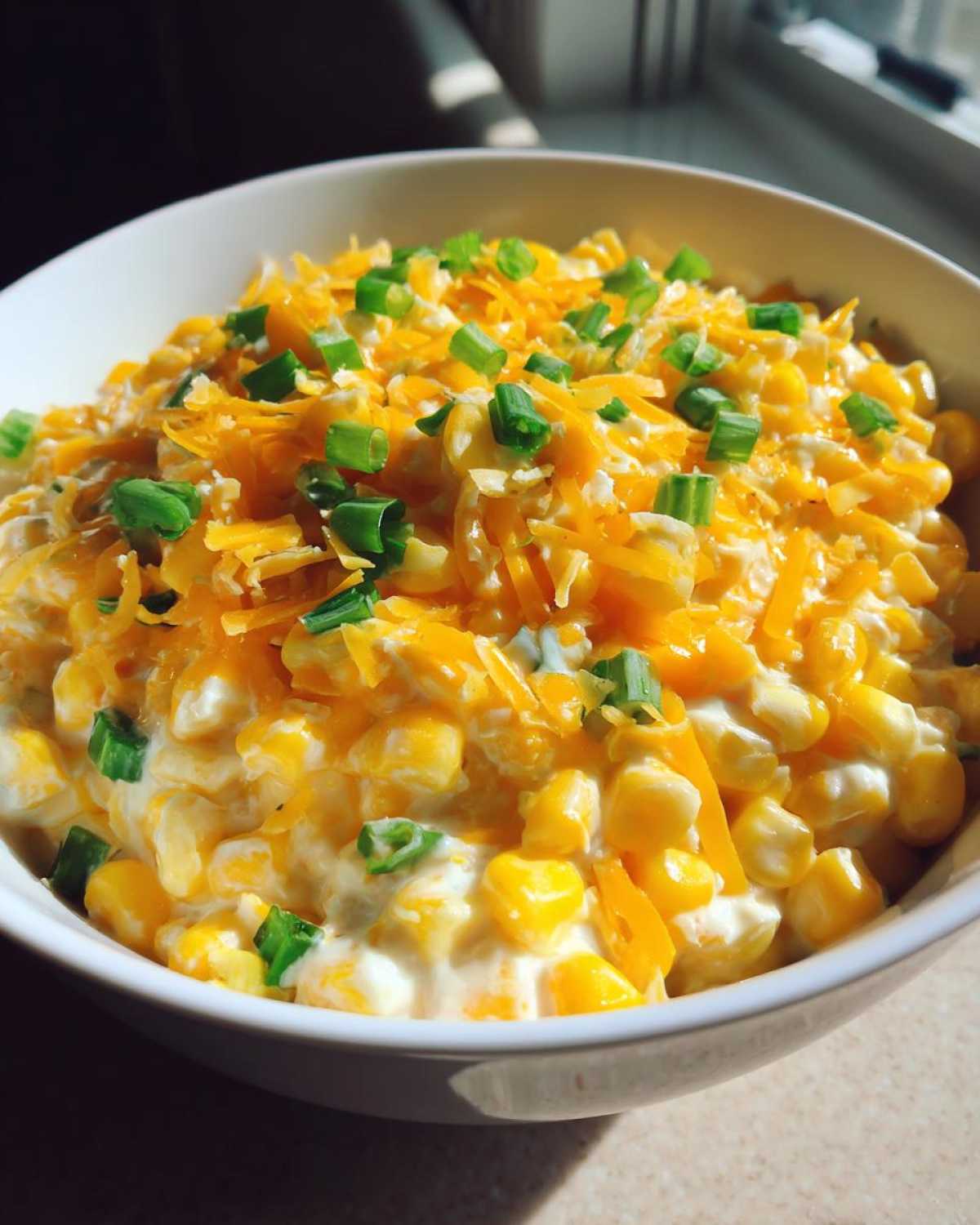 Close-up of a white bowl filled with creamy corn dip with sour cream, topped with shredded cheddar cheese and chopped green onions.
