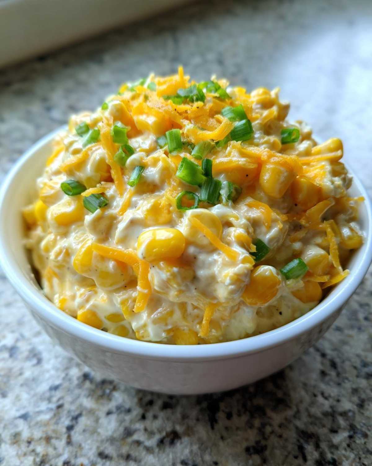 A white bowl filled with creamy corn dip with sour cream, topped with shredded cheddar cheese and chopped green onions.