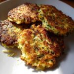Close-up of several golden brown zucchini fritters on a white plate, showing crispy edges and flecks of green zucchini.