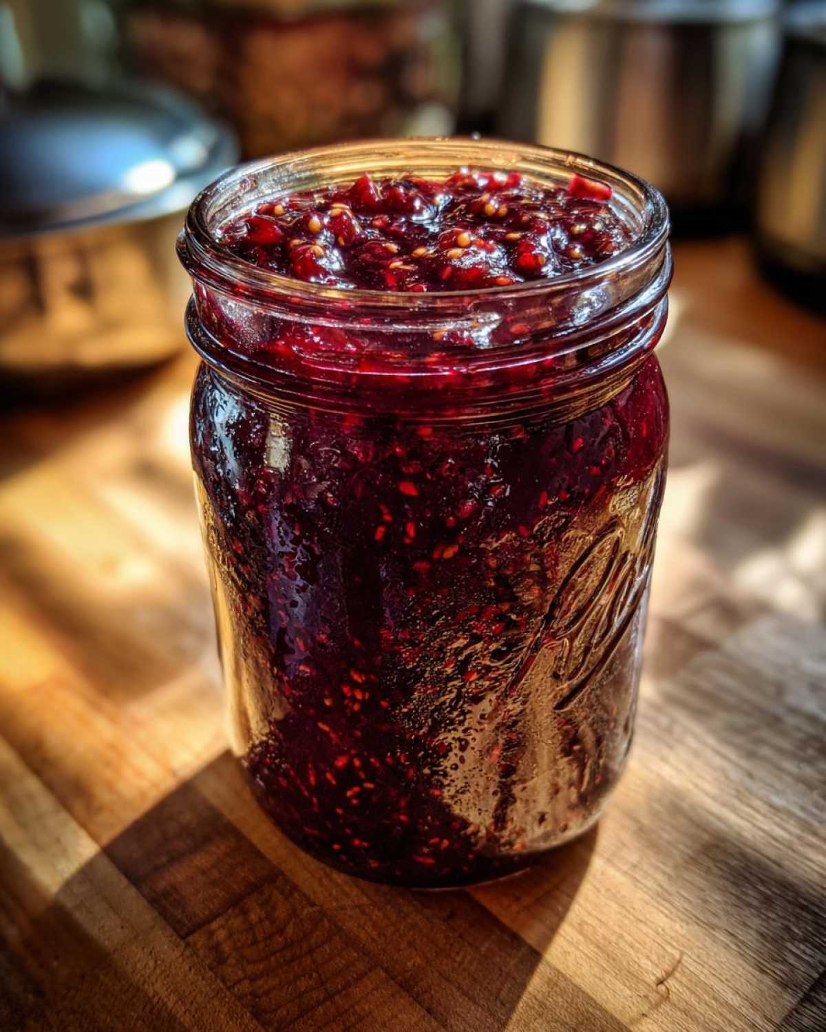 A close-up of a glass mason jar filled with dark red, glistening fig jam on a wooden surface.
