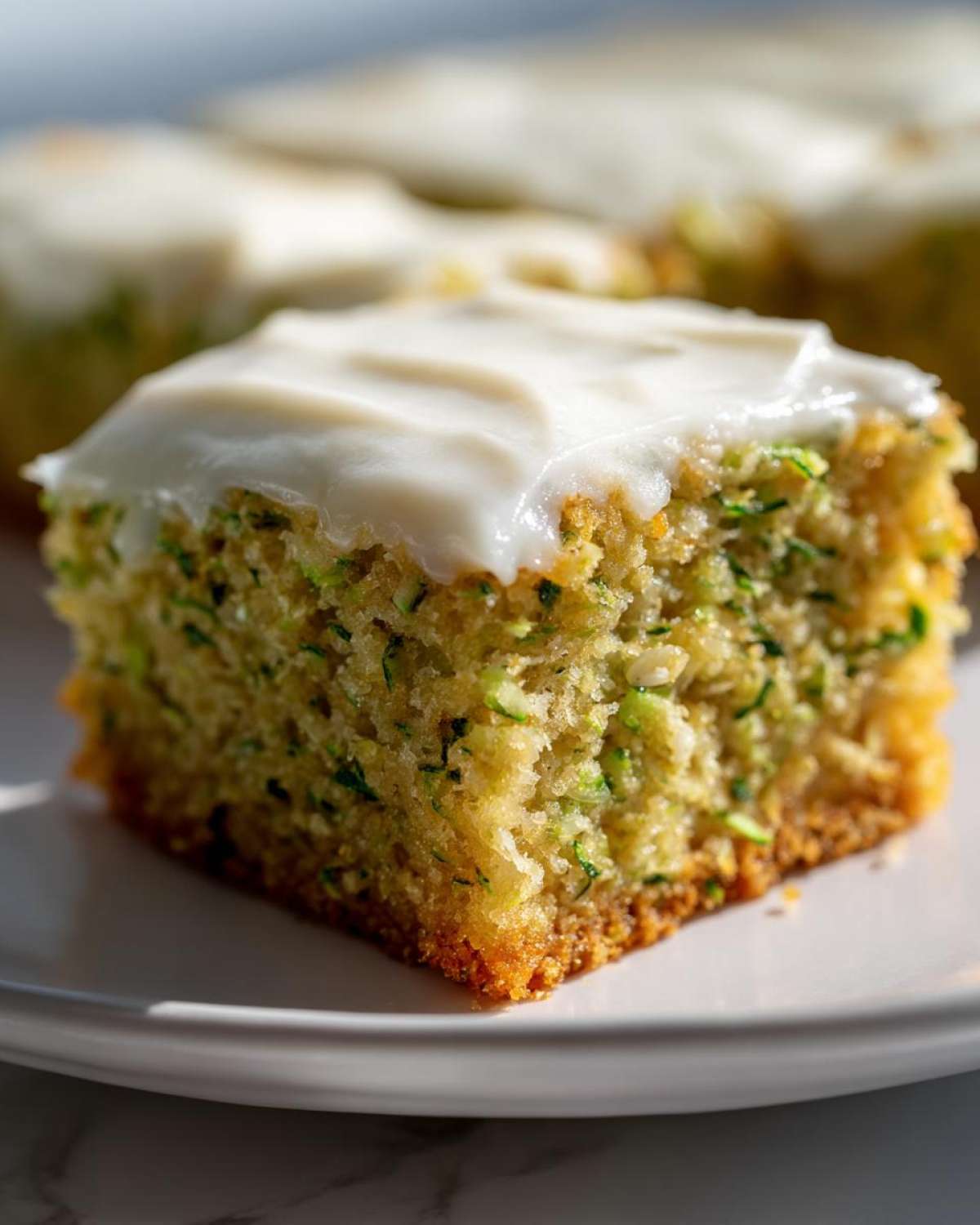 A close-up shot of a single square zucchini bar with white frosting on a white plate.