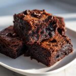 Close-up of a stack of rich, dark zucchini brownies topped with chocolate chips on a white plate.