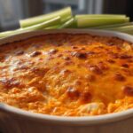 Close-up of a bubbling, golden-brown high protein buffalo chicken dip in a white baking dish, with celery sticks in the background.
