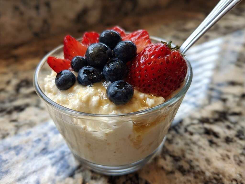 Close-up of a glass bowl filled with creamy high protein overnight oats, topped with fresh blueberries, sliced strawberries, and a drizzle of honey. A spoon is visible on the right.