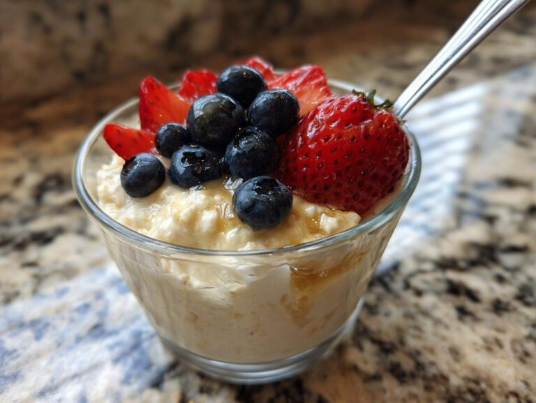 Close-up of a glass bowl filled with creamy high protein overnight oats, topped with fresh blueberries, sliced strawberries, and a drizzle of honey. A spoon is visible on the right.