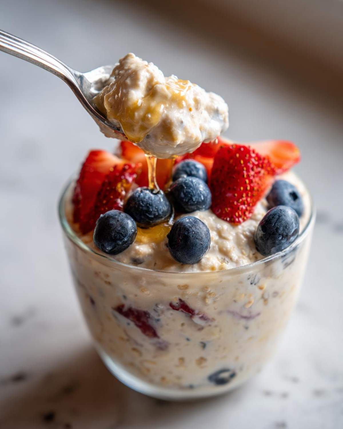 A spoonful of high protein overnight oats being lifted from a glass, topped with fresh blueberries, sliced strawberries, and a drizzle of honey.