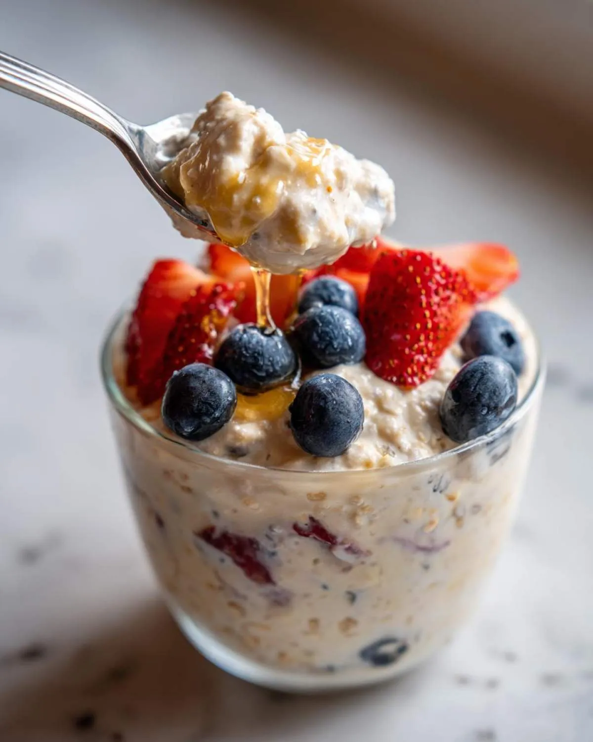 A spoonful of high protein overnight oats being lifted from a glass, topped with fresh blueberries, sliced strawberries, and a drizzle of honey.