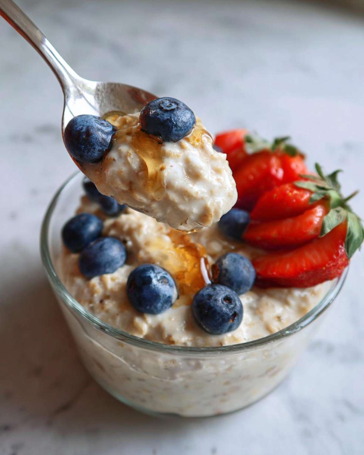 A spoonful of high protein overnight oats topped with blueberries and honey, with a bowl of oats and strawberries in the background.
