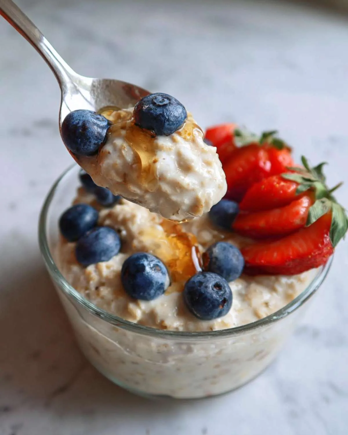A spoonful of high protein overnight oats topped with blueberries and honey, with a bowl of oats and strawberries in the background.
