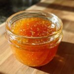 A small glass jar filled with bright orange apricot jam sits on a wooden surface in sunlight.