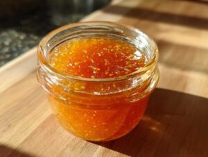 A small glass jar filled with bright orange apricot jam sits on a wooden surface in sunlight.