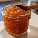 Close-up of a glass jar filled with homemade peach jam, with a wooden spoon resting on top.