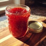 A clear glass jar filled with vibrant red strawberry jam, sitting on a wooden cutting board next to its golden lid.