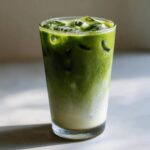 A tall glass filled with a vibrant green iced matcha latte, showing the layers of milk and matcha with ice cubes.