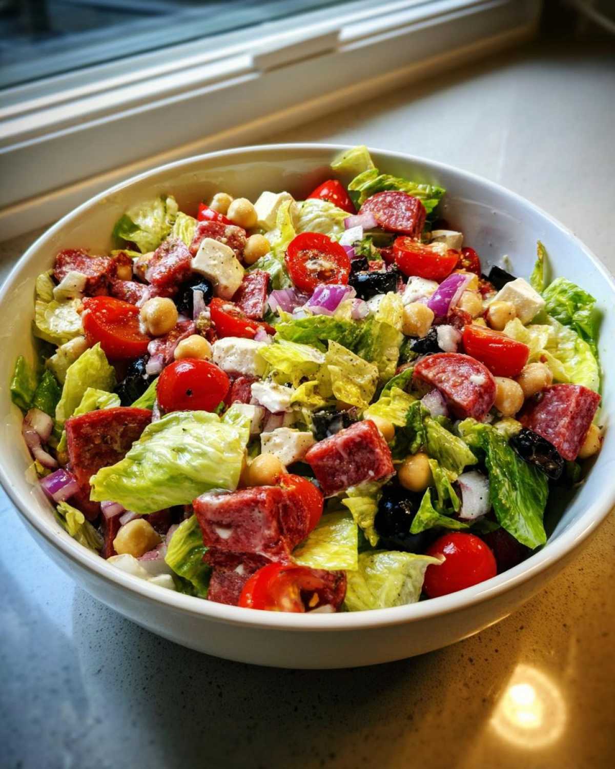 A close-up view of a fresh Italian chopped salad in a white bowl, featuring lettuce, cherry tomatoes, salami, chickpeas, and cheese.