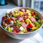 A large white bowl filled with Italian chopped salad, featuring lettuce, salami, cheese, chickpeas, olives, and red onion.