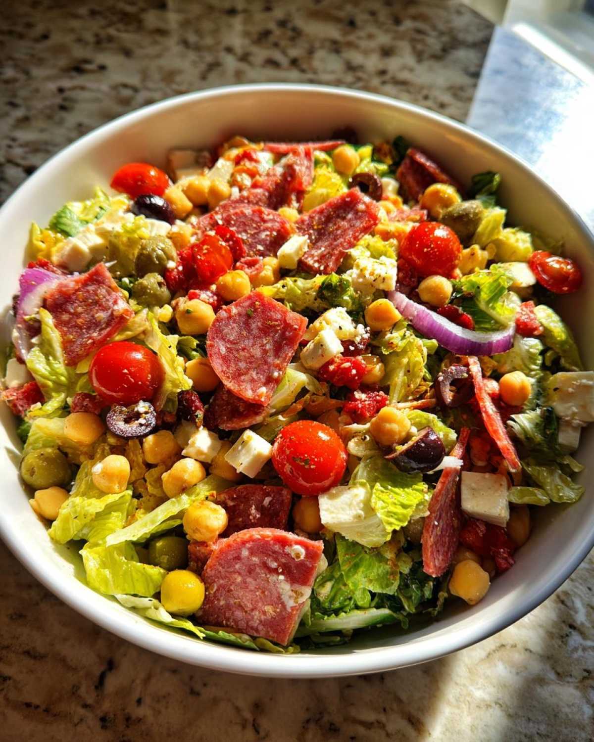 A close-up, top-down view of a colorful Italian chopped salad in a white bowl, featuring salami, tomatoes, and chickpeas.