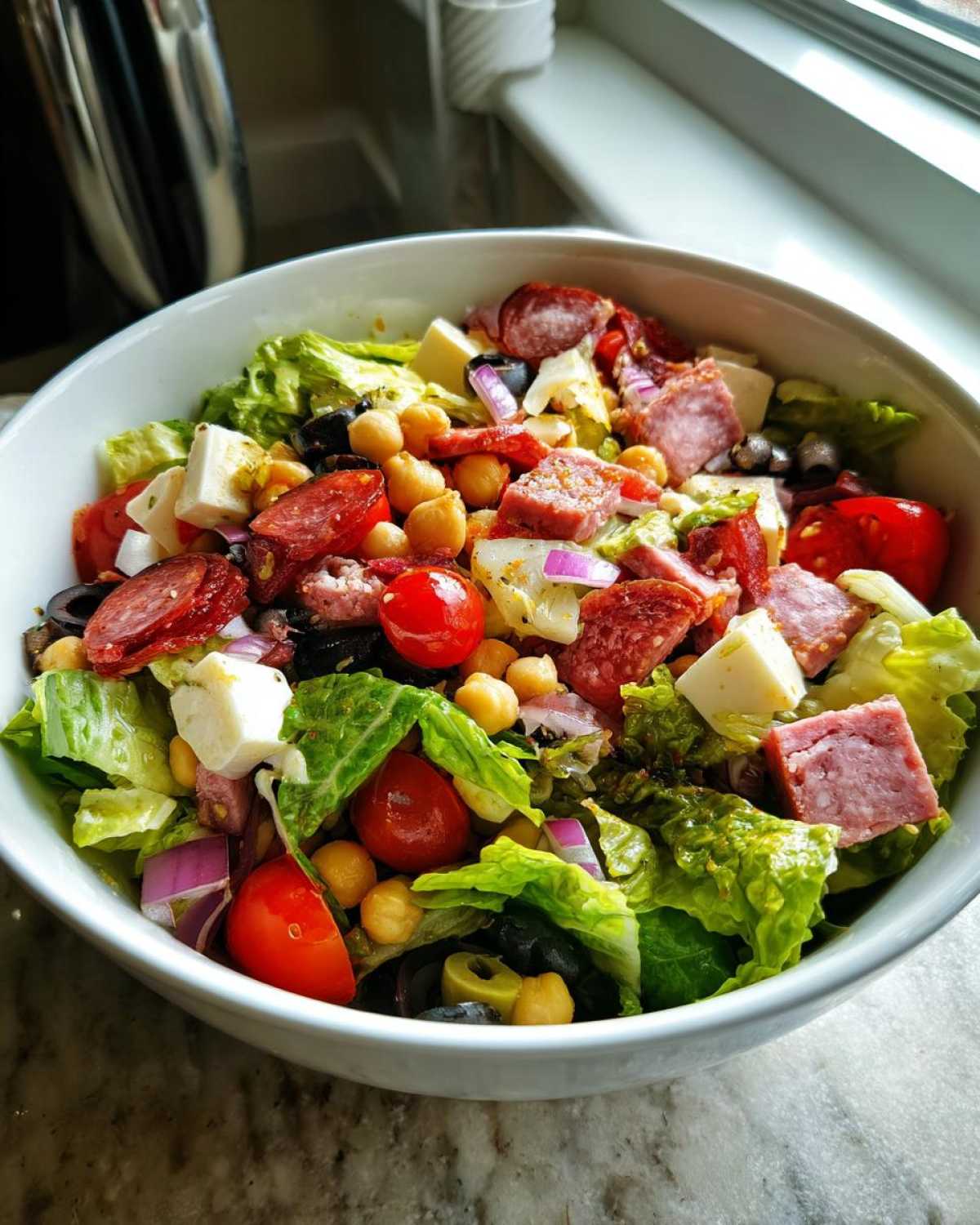 A close-up of a colorful Italian chopped salad in a white bowl, featuring lettuce, tomatoes, chickpeas, salami, and cheese.